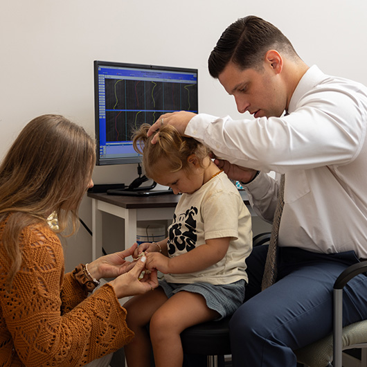 Dr. Zachary Shaw using the natural effective alternative to heal a patient. A chiropractor performing an upper cervical spine assessment on a child, while the child's mother assists during the examination. A computer monitor displaying spinal data is visible in the background.